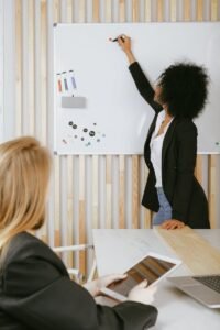 Deux femmes participent à une présentation commerciale avec un tableau blanc dans un bureau moderne.
