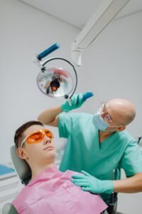 Un dentiste en tenue verte examine un patient portant des lunettes de protection sous une lumière dentaire.