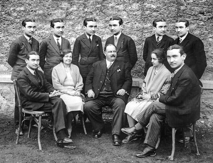 Groupe de personnes en tenue formelle, assis et debout sur des chaises, devant un mur de pierre Groupe de personnes en tenue formelle, assis et debout sur des chaises, devant un mur de pierre